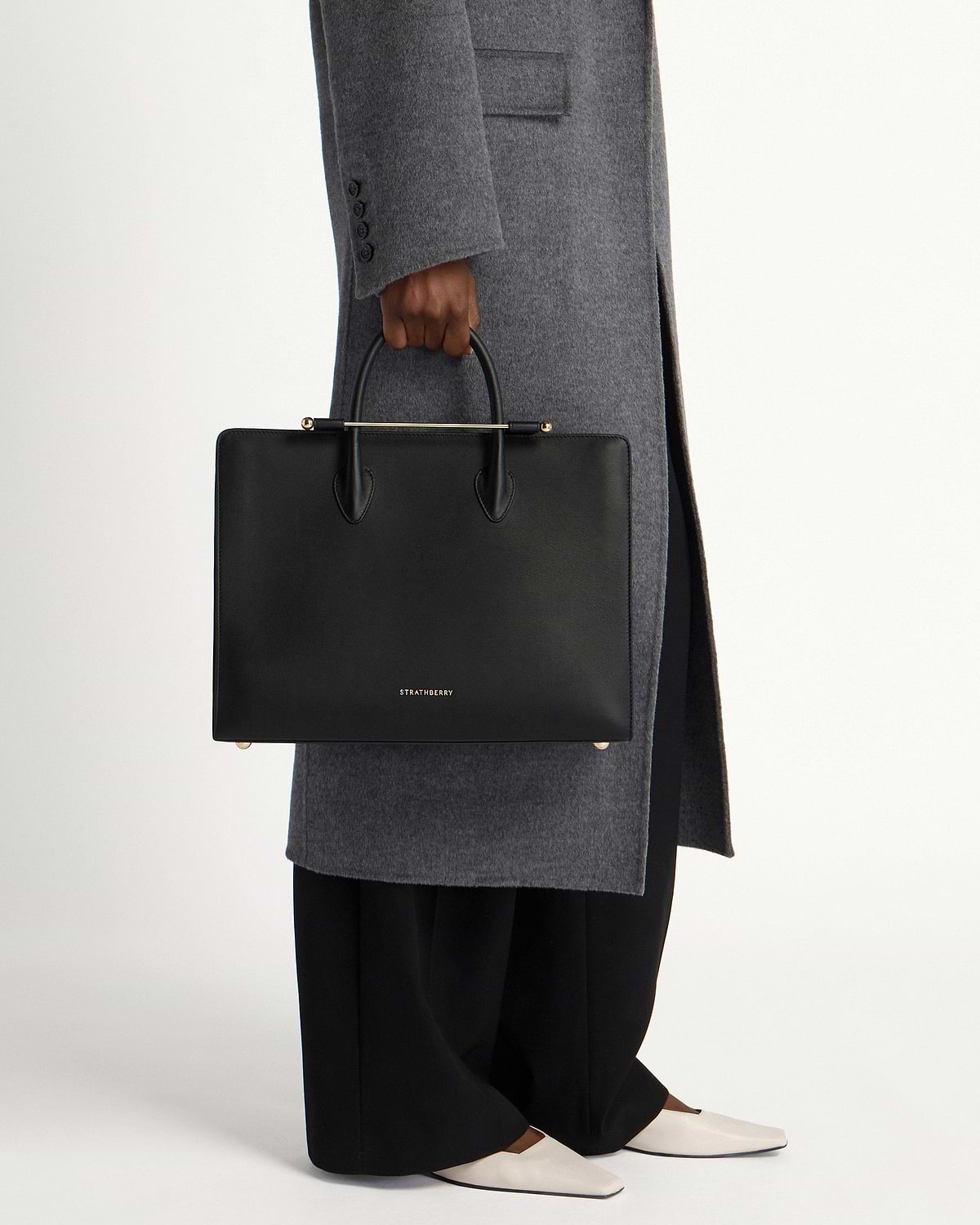 Black Strathberry leather tote with top handles held by model wearing grey coat