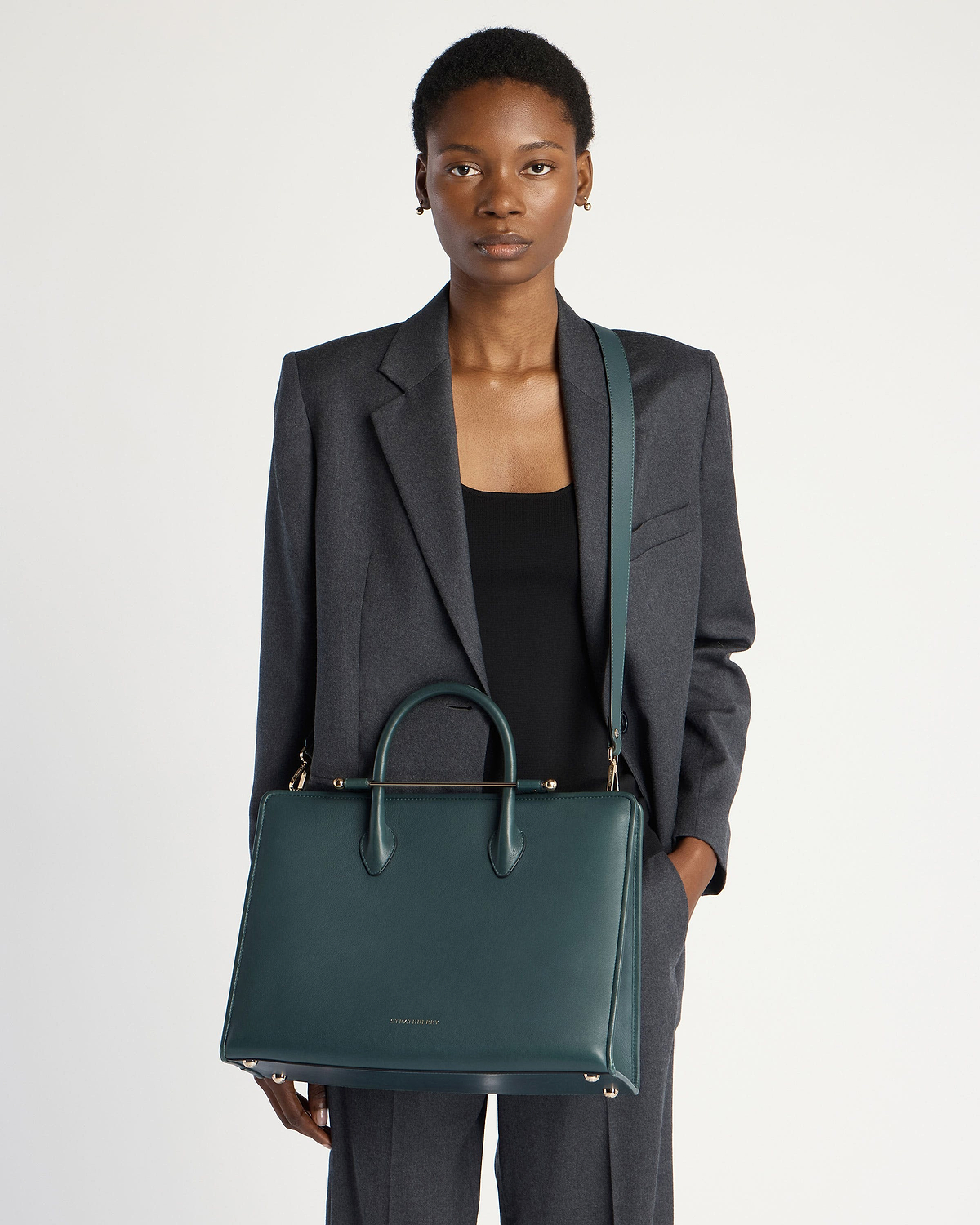 Model wearing a grey blazer holds a bottle-green Strathberry leather tote bag.