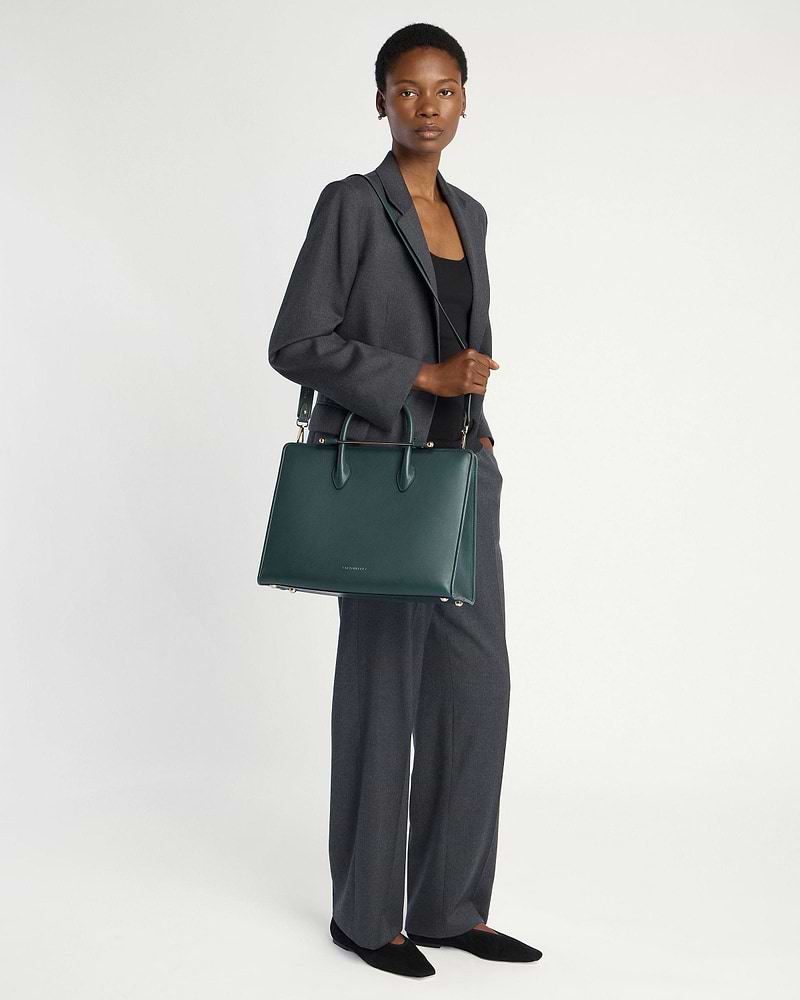 Woman in grey suit standing, holding Strathberry bottle green tote bag with strap.