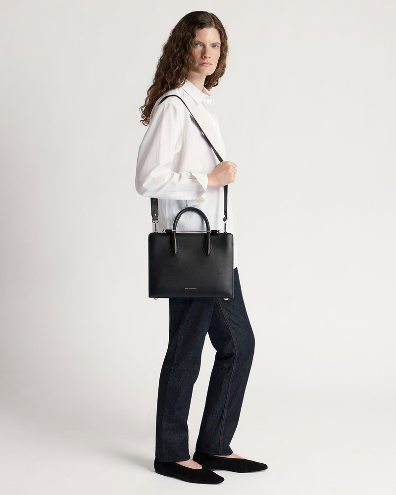Model wearing a white shirt and jeans carrying a black Strathberry midi tote.