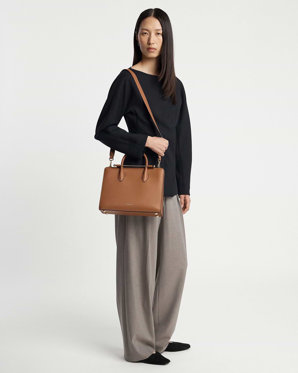 Model wearing black top and grey wide trousers holding chestnut midi tote bag.