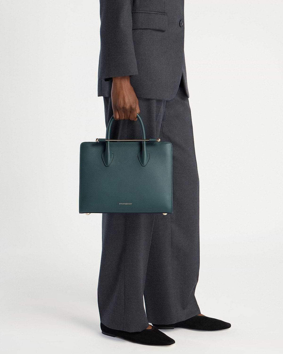 Model wearing grey suit holding bottle green Strathberry midi tote at side
