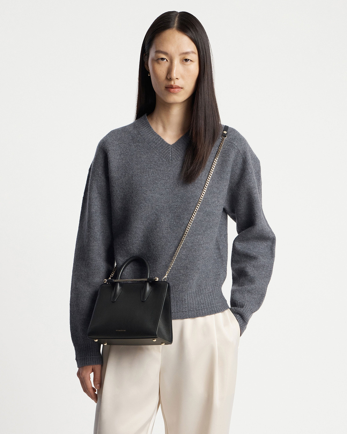 Model wearing grey sweater and cream trousers carrying a black mini tote bag