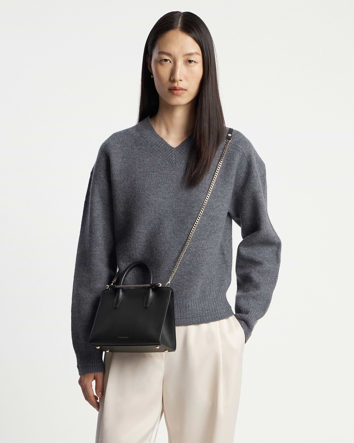 Model wearing grey sweater and cream trousers carrying a black mini tote bag