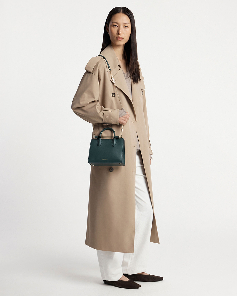 Woman in beige trench coat holding bottle green mini tote with chain shoulder strap.