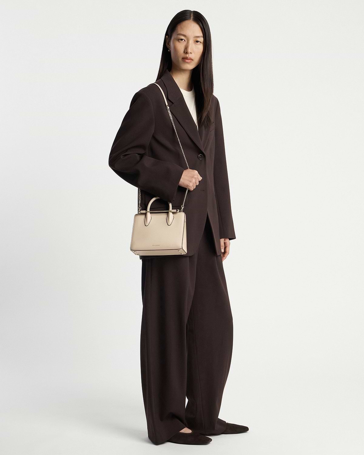 Model in dark brown suit holding oat mini tote by top handles and chain strap.