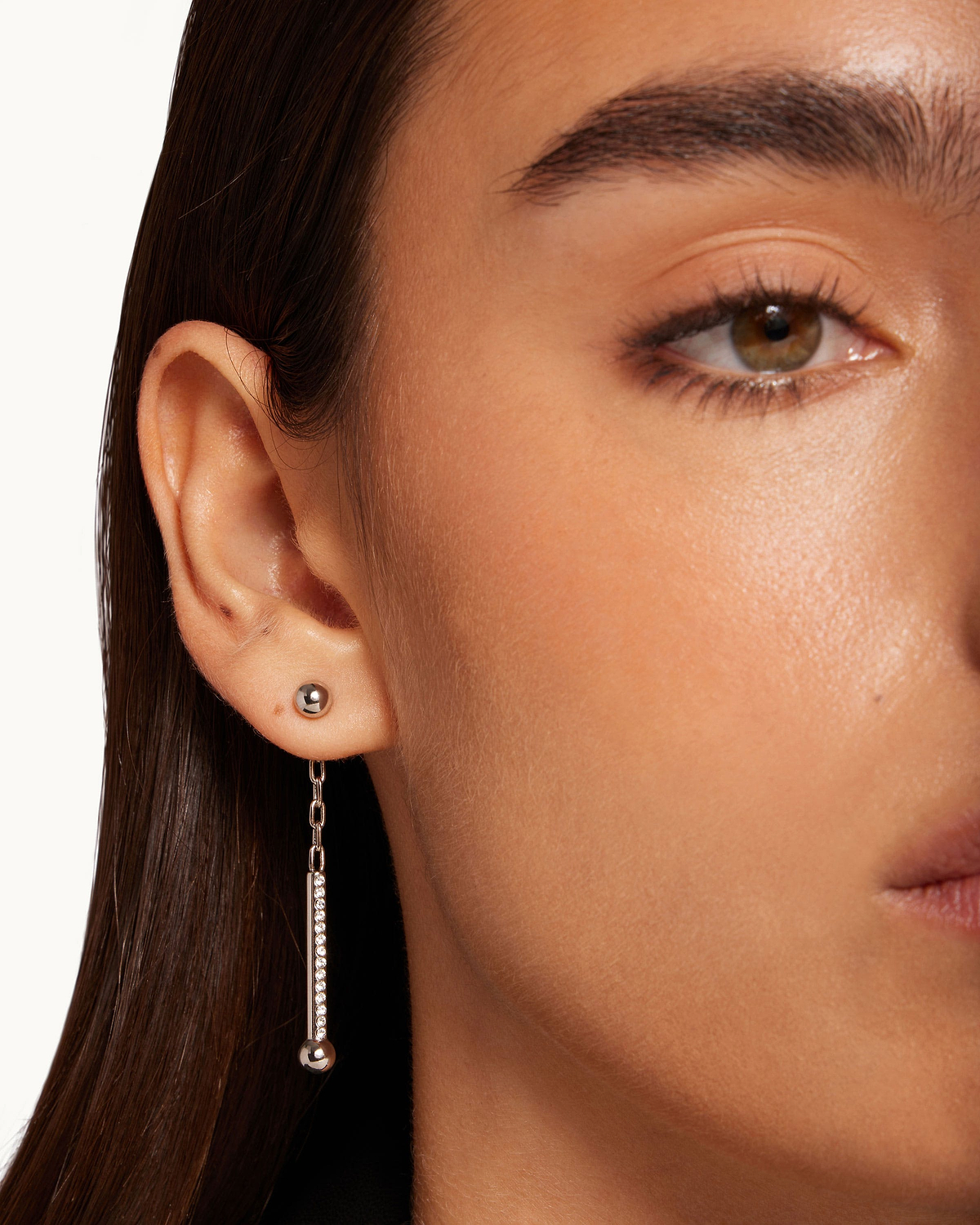 Woman's ear with silver drop earring featuring crystal bar and metal ball; small stud above.