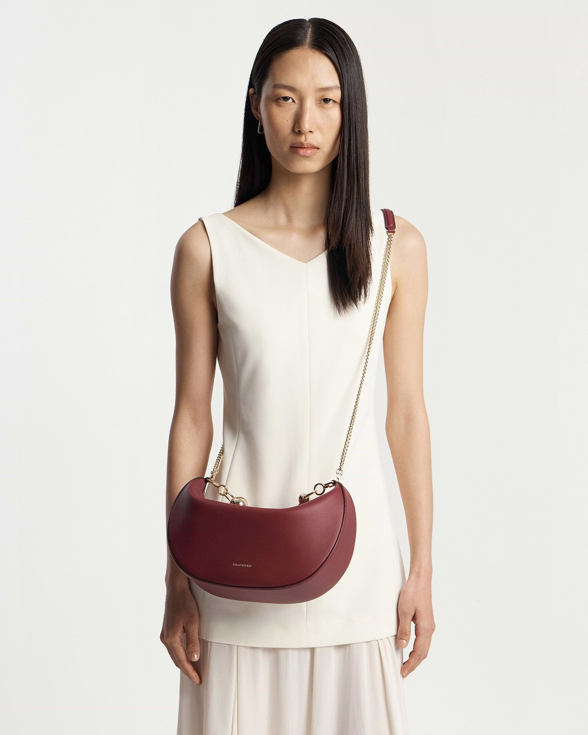 Model wearing white sleeveless dress carrying oxblood crescent moon bag with gold chain strap.