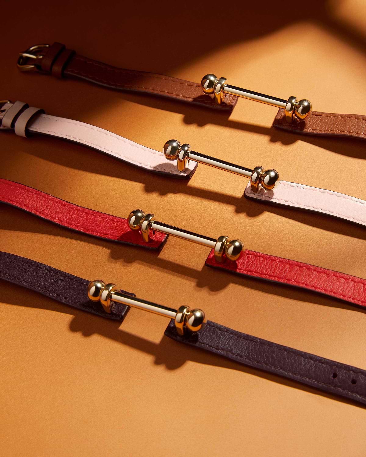 Four leather bracelets with 22 carat gold-gilded bar hardware on a warm golden background.