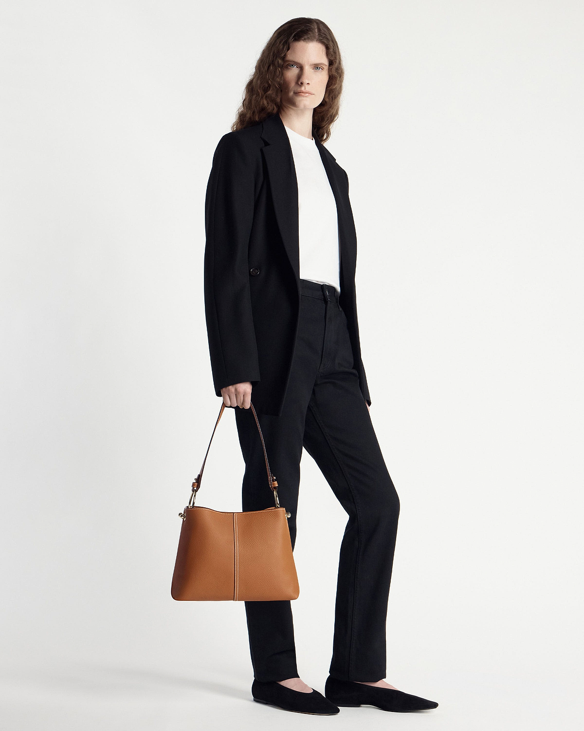 Model in a black blazer and trousers holding a tan hobo bag at her side.