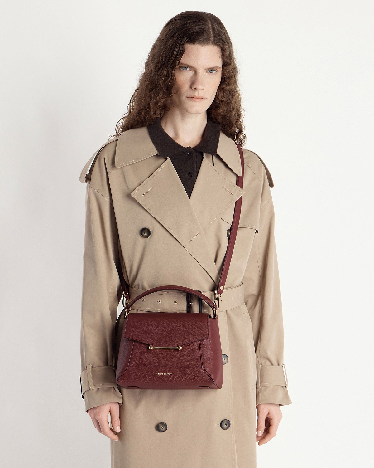 Model wearing a beige trench coat carrying an oxblood mosaic bag by Strathberry crossbody.