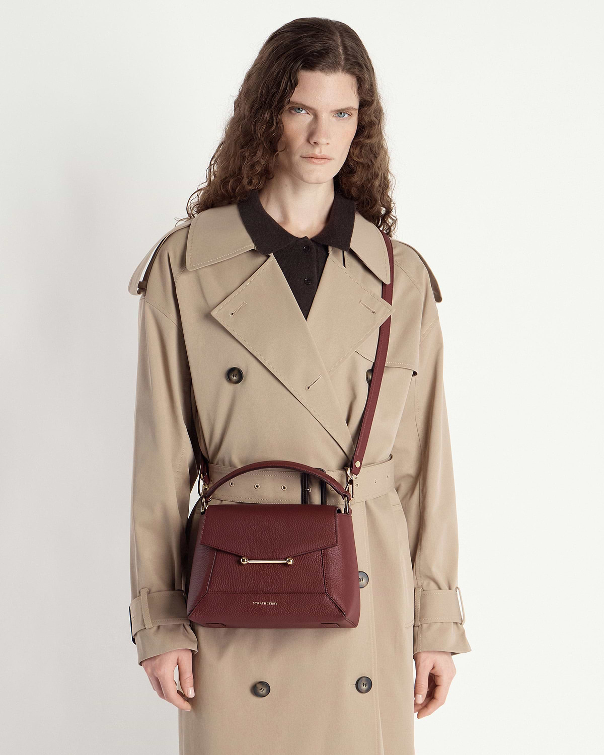 Model wearing a beige trench coat carrying an oxblood mosaic bag by Strathberry crossbody.