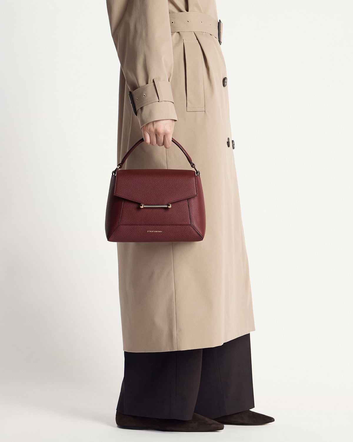 Model carrying oxblood Strathberry Mosaic bag with beige trench coat and black trousers.