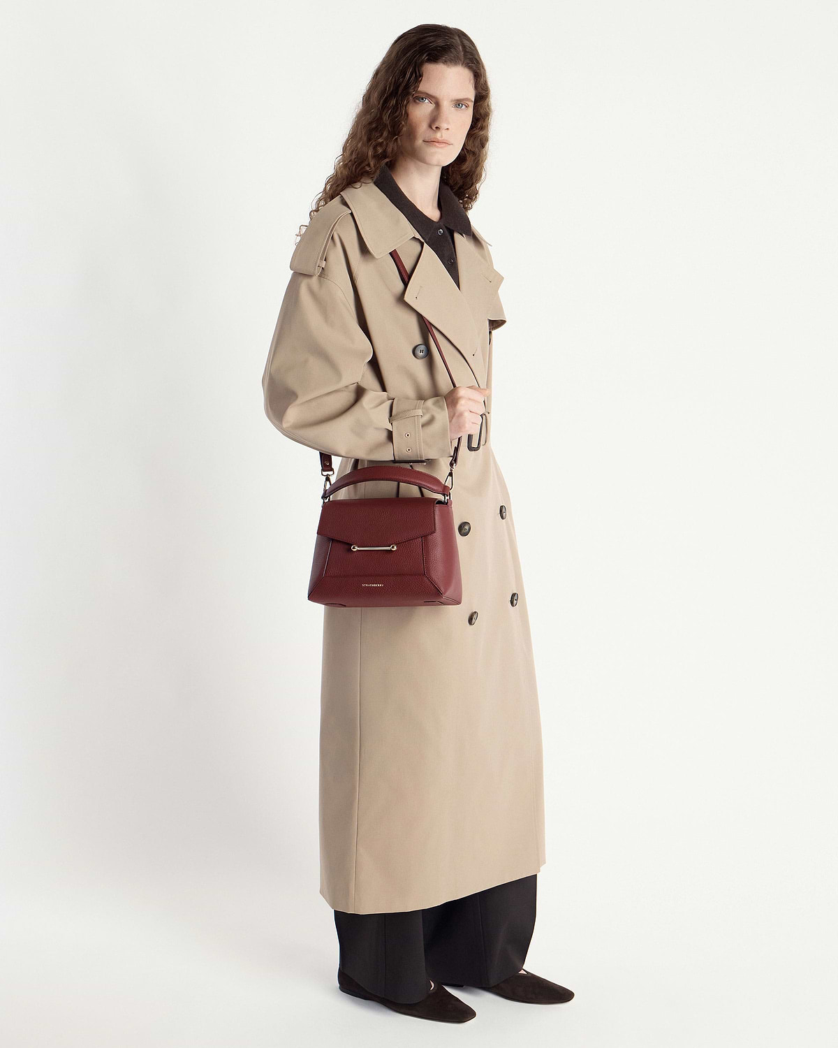 Model in beige trench coat holding an oxblood mosaic bag by the handle.