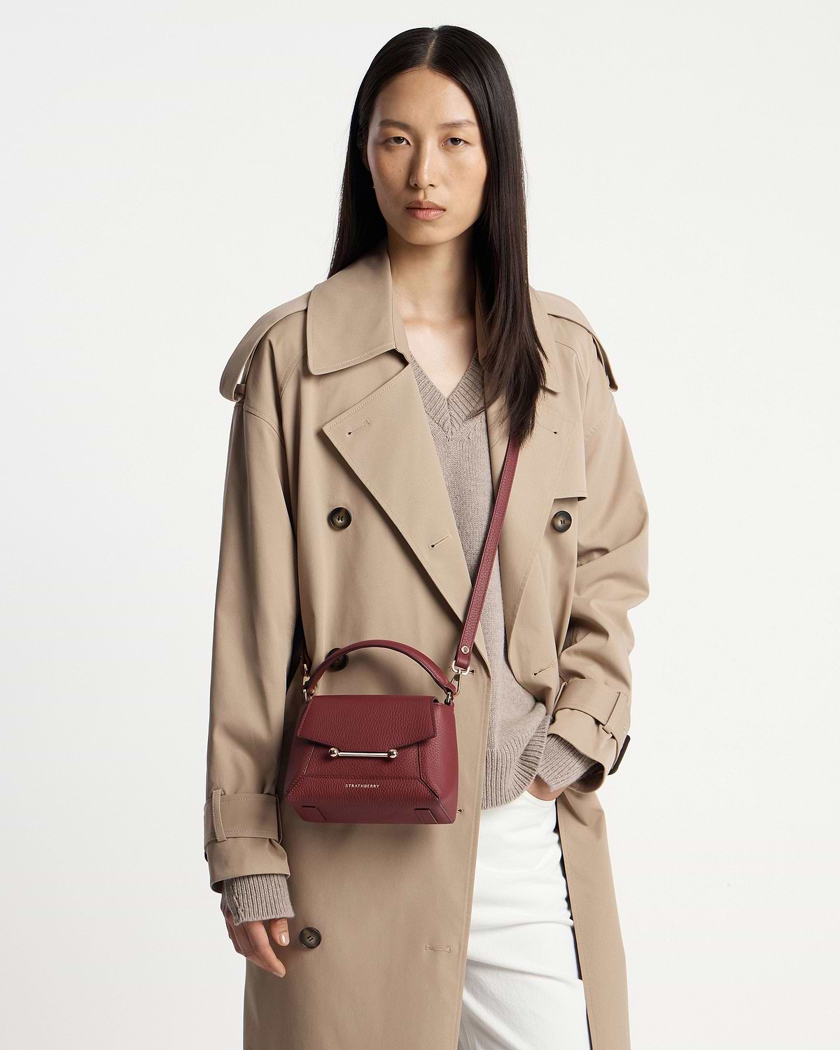 Model wearing beige trench coat carrying Strathberry Mosaic Nano bag in oxblood crossbody.