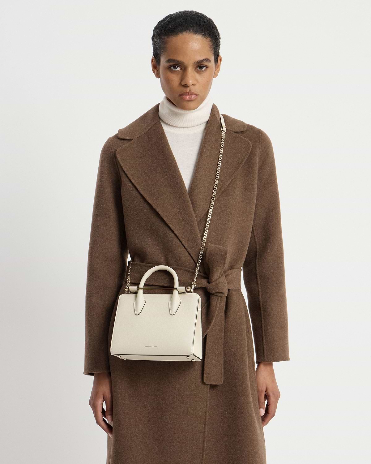 Model wearing brown coat with vanilla Strathberry mini tote on gold chain.
