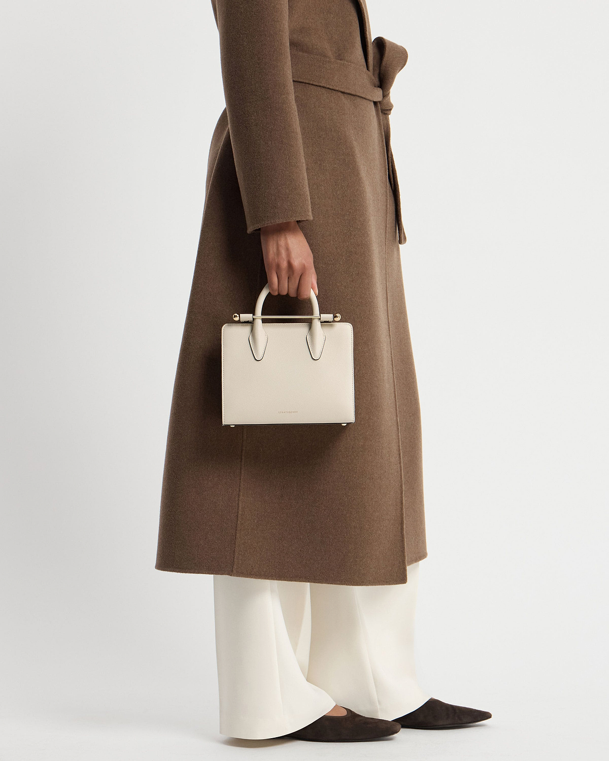 Model cropped below waist wearing brown coat, holding vanilla mini tote by top handles.