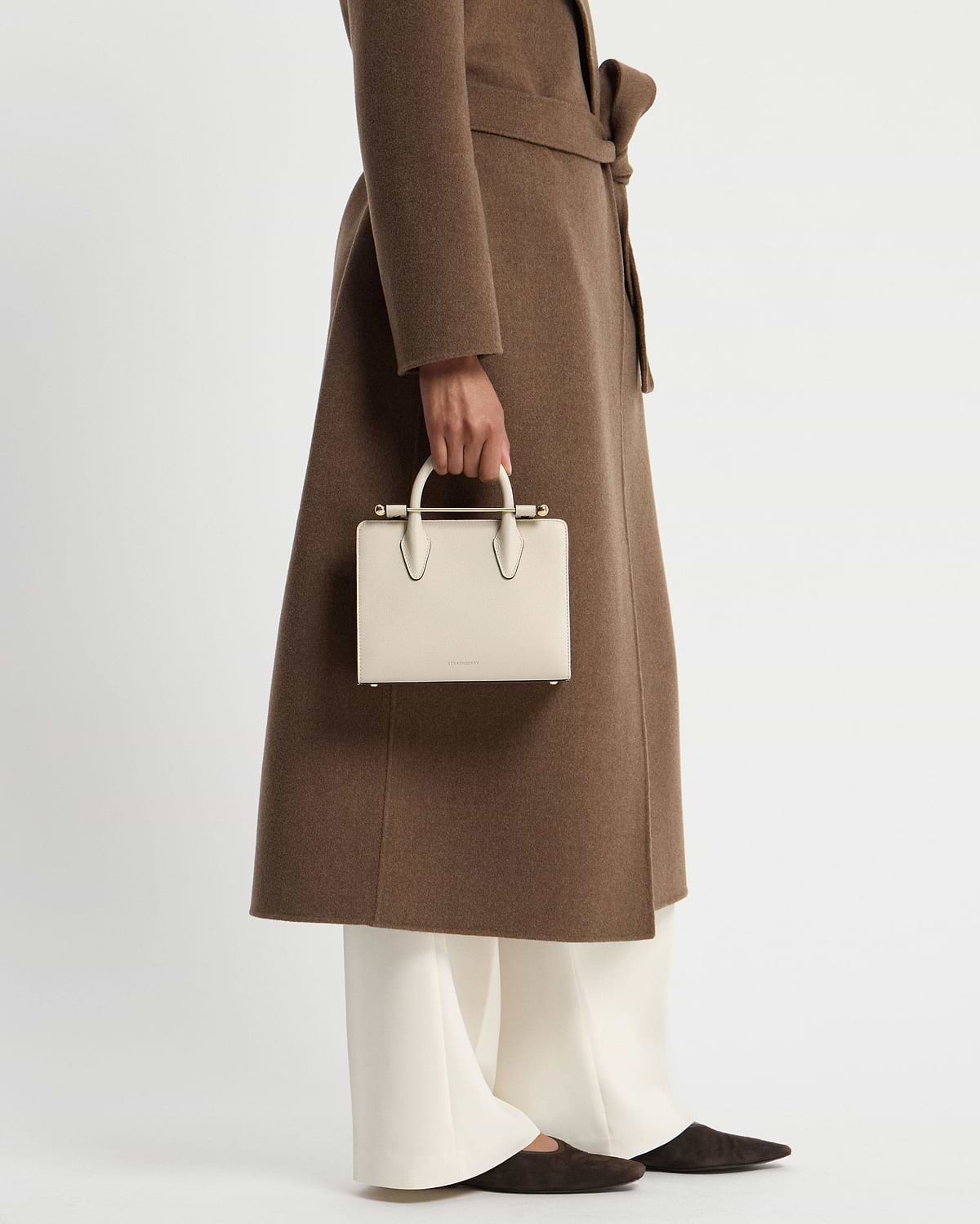 Model cropped below waist wearing brown coat, holding vanilla mini tote by top handles.