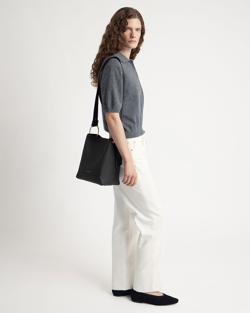 Model wearing grey short-sleeve polo and white jeans carrying black bucket bag over shoulder