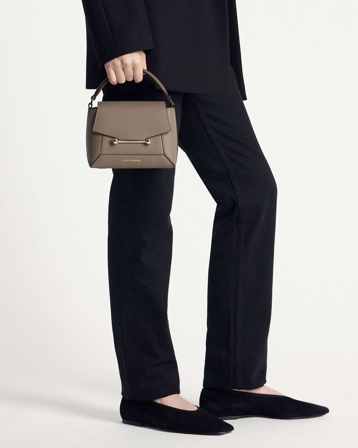 Model waist-down holding taupe Strathberry Mosaic Nano top-handle bag with gold bar closure.