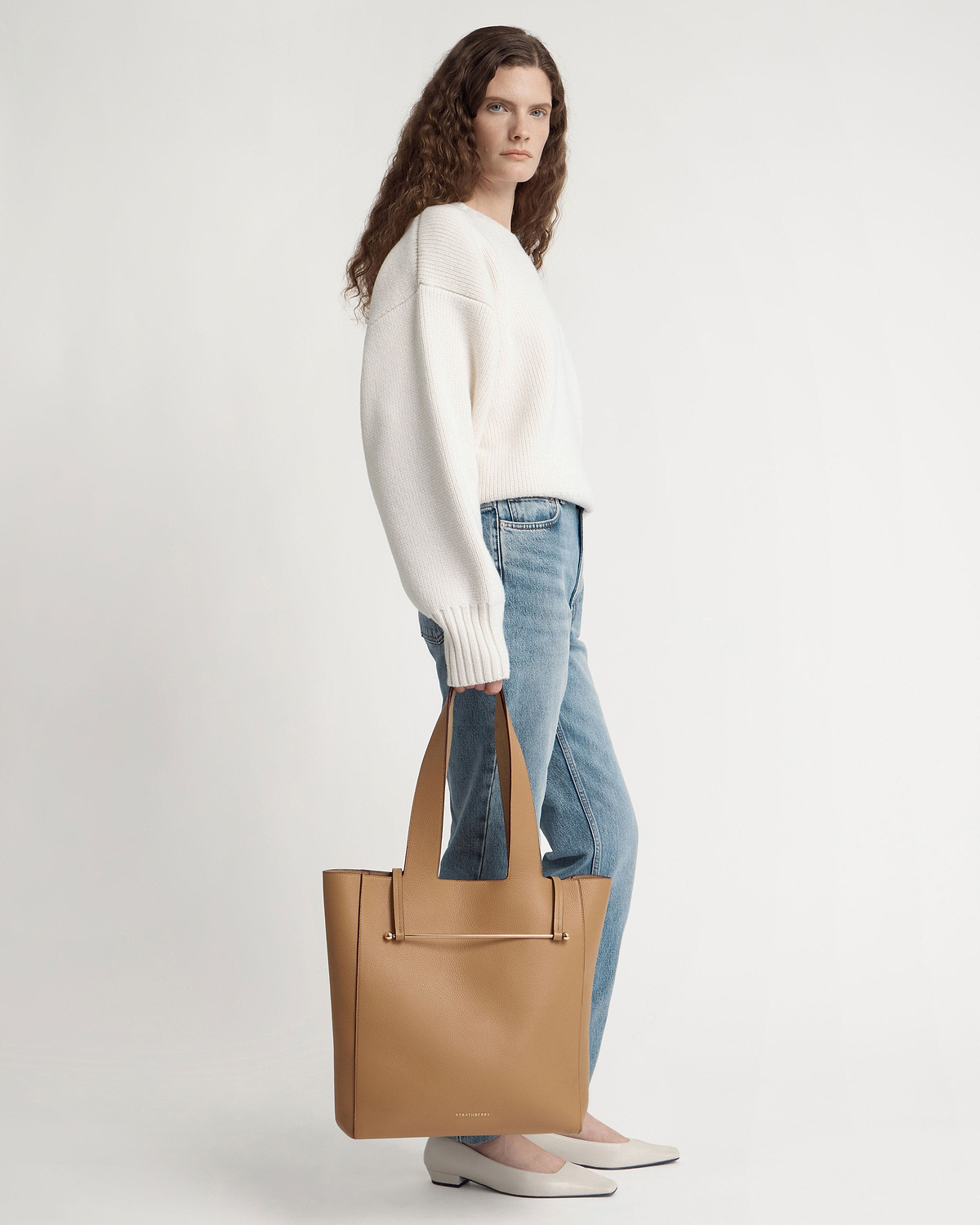 Woman standing in white jumper and jeans holding a caramel tote bag