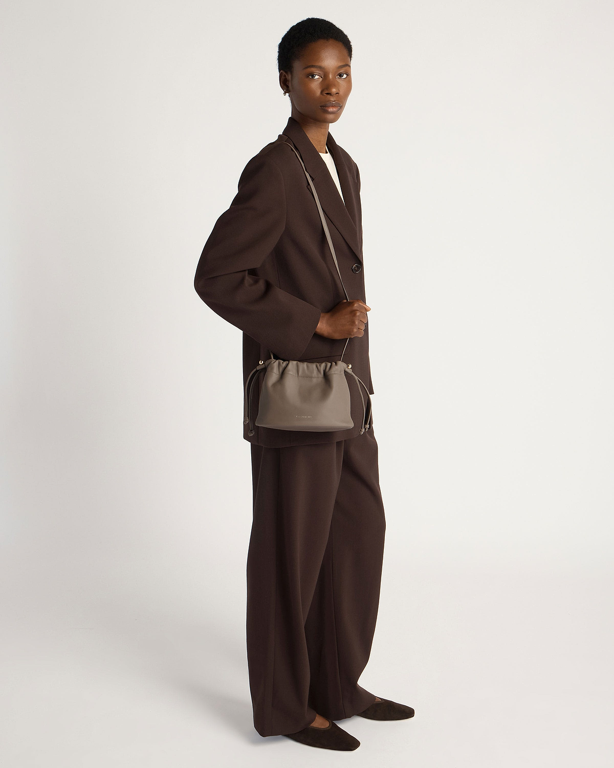 Model in dark brown suit carrying a taupe drawstring cross-body bag.