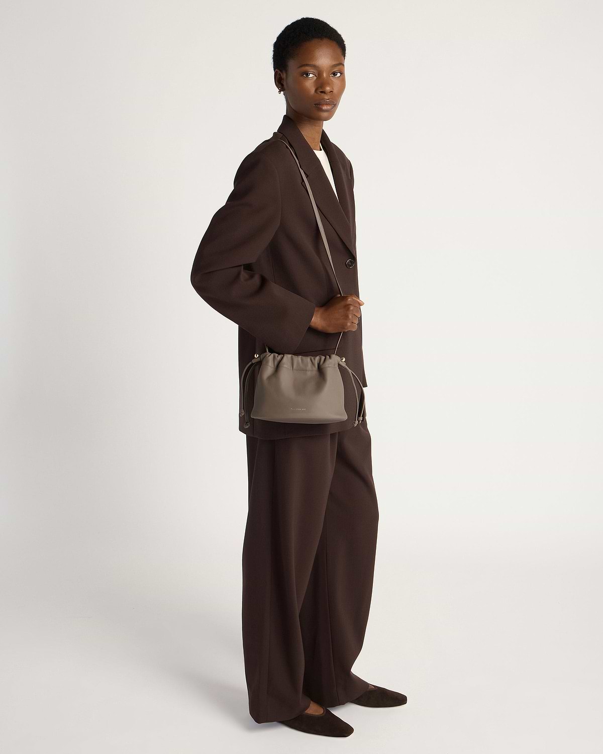 Model in dark brown suit carrying a taupe drawstring cross-body bag.