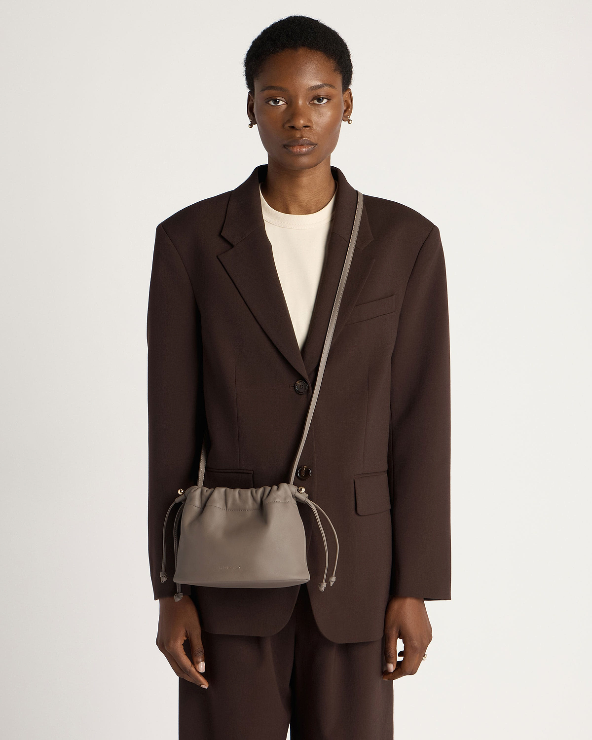 Model wearing a taupe drawstring bag crossbody over a brown blazer and trousers