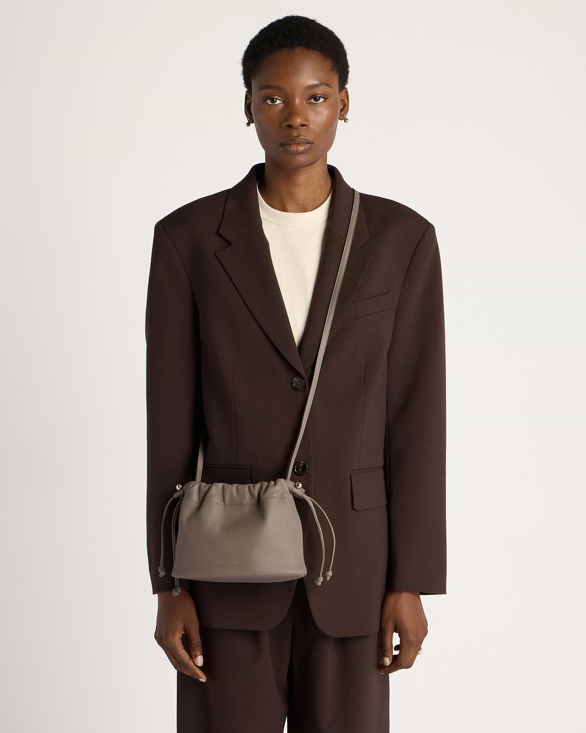 Model wearing a taupe drawstring bag crossbody over a brown blazer and trousers