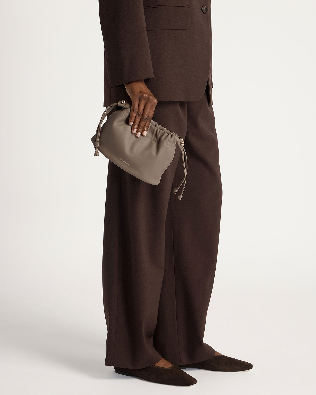 Taupe drawstring bag held by model wearing a brown suit and flats.