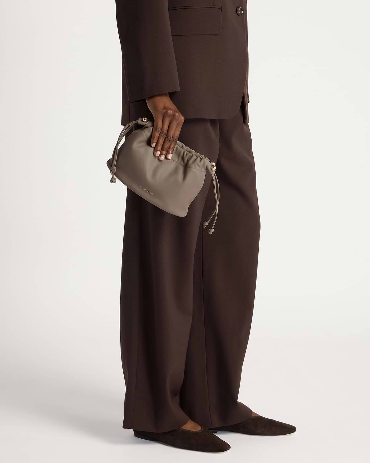 Taupe drawstring bag held by model wearing a brown suit and flats.
