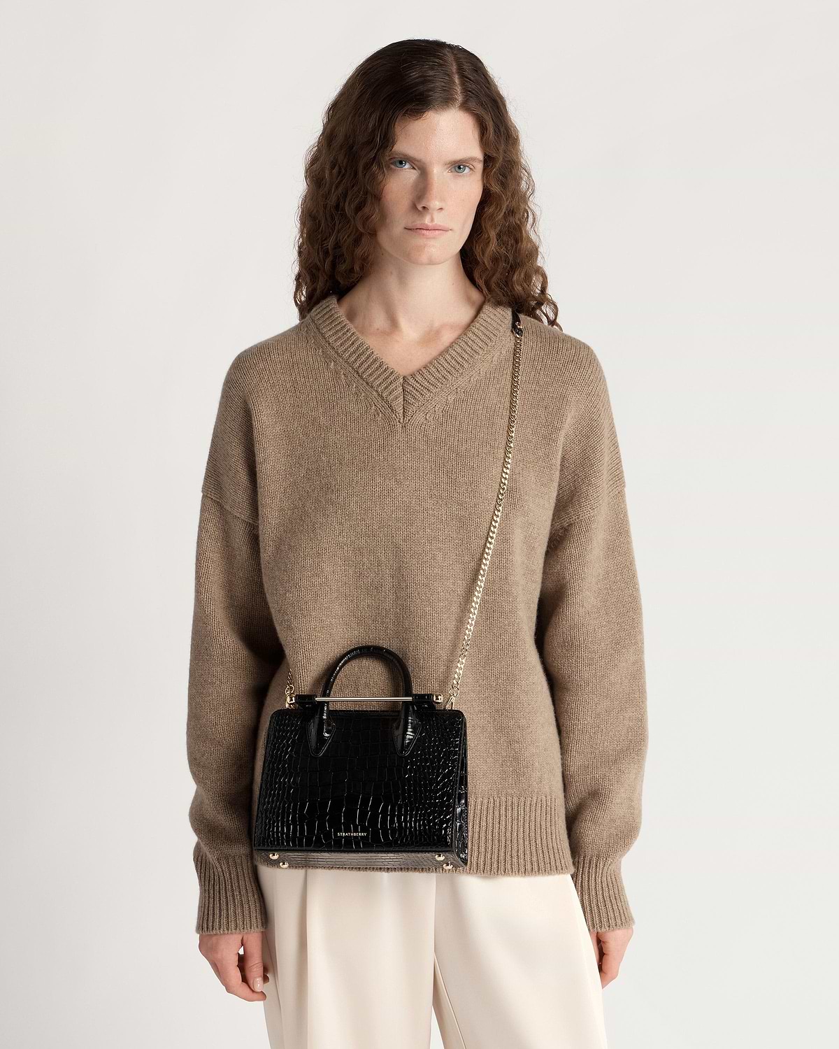 Woman wearing beige jumper carrying black croc Strathberry mini tote on a chain