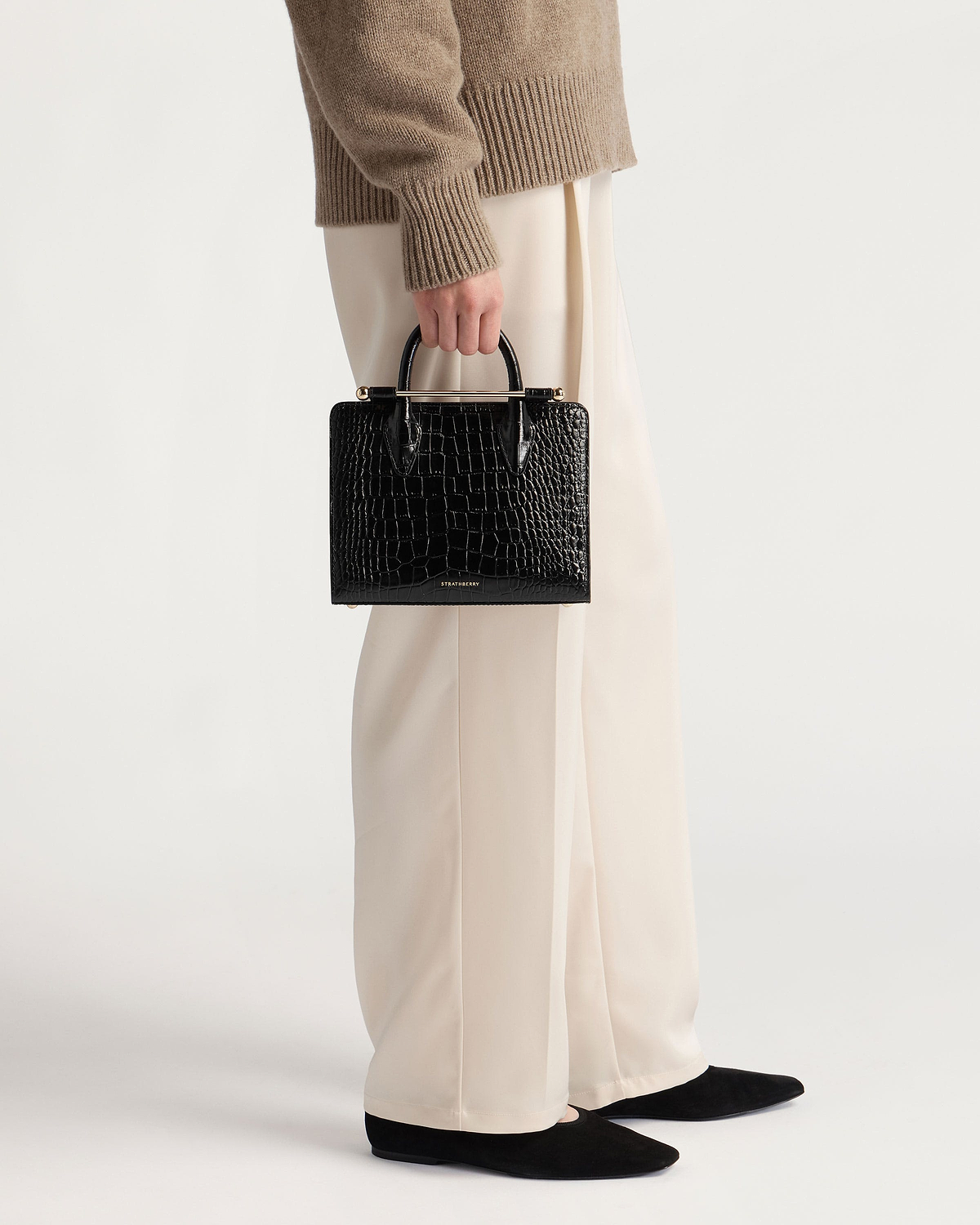 Hand holding black croc mini tote by Strathberry against cream wide-leg trousers.