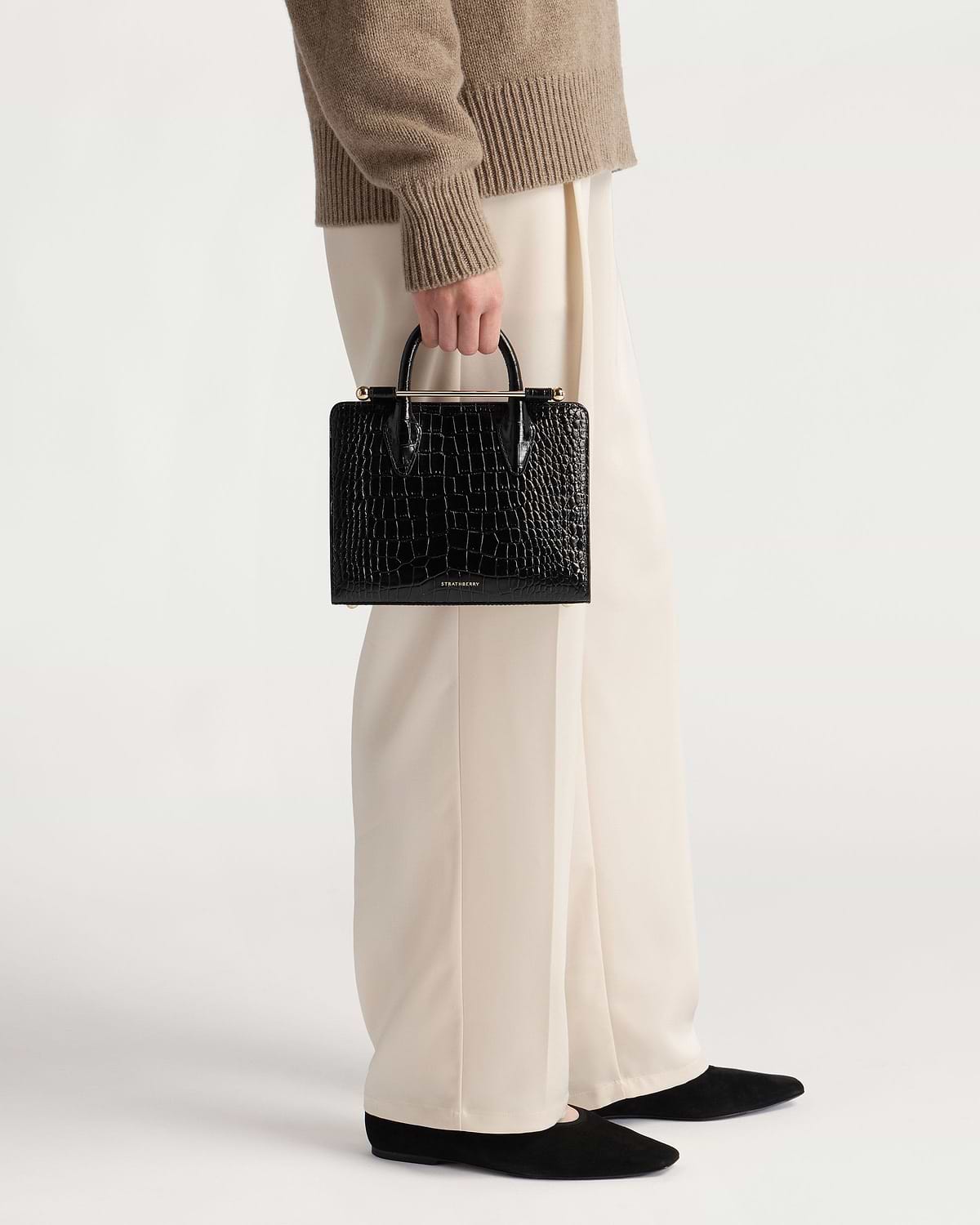 Hand holding black croc mini tote by Strathberry against cream wide-leg trousers.