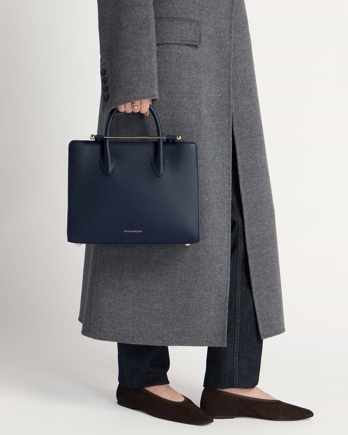 A hand holds Strathberry marine blue midi tote against grey coat and jeans.
