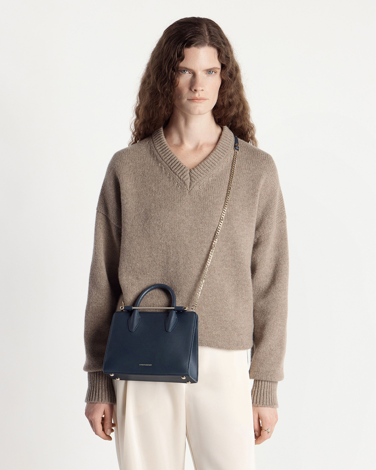 Model wearing beige jumper with marine blue Strathberry mini tote and chain strap.