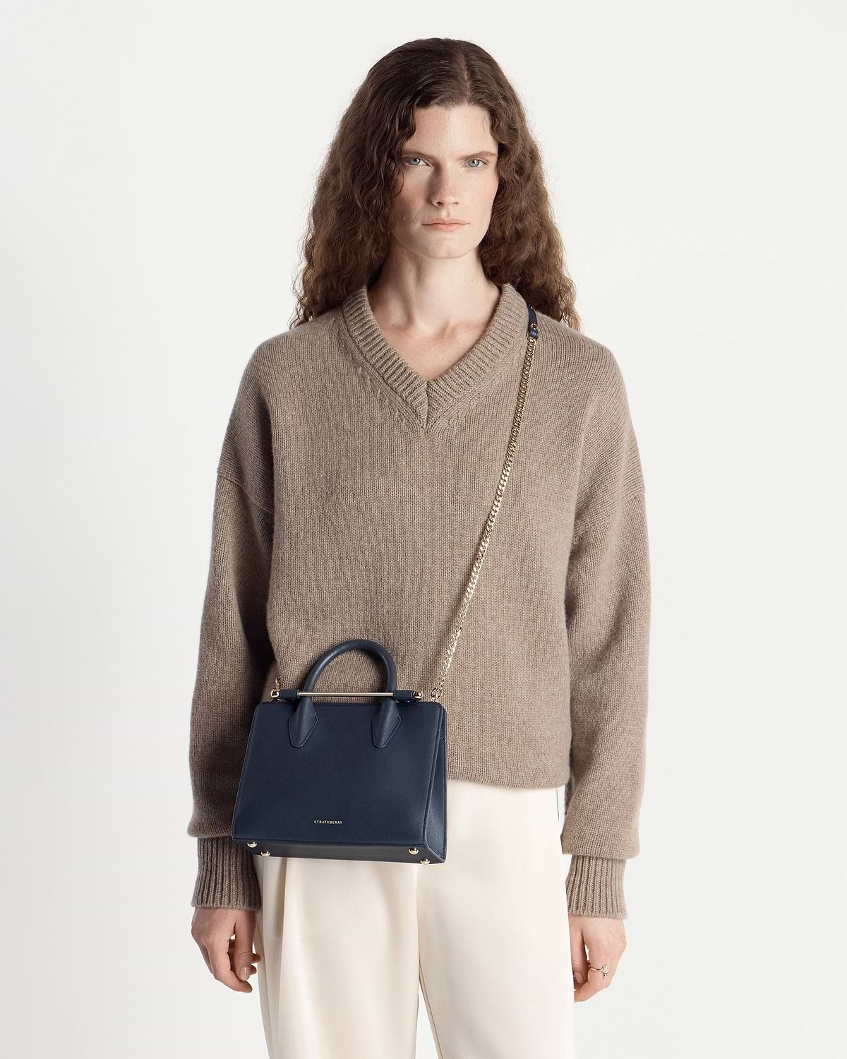 Model wearing beige jumper with marine blue Strathberry mini tote and chain strap.