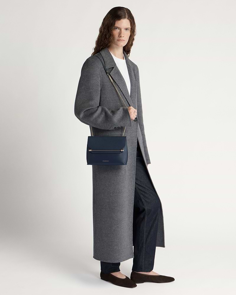 Model wearing long grey coat carrying marine blue shoulder bag with gold bar detail.