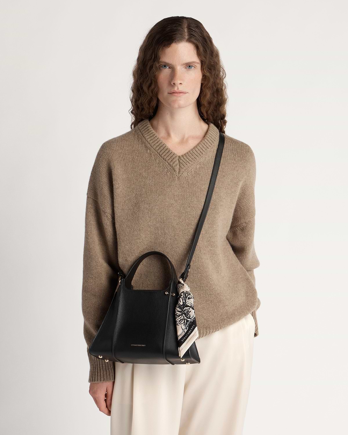 Model wearing beige jumper carrying black Strathberry Arkshell bag with shoulder strap and scarf.