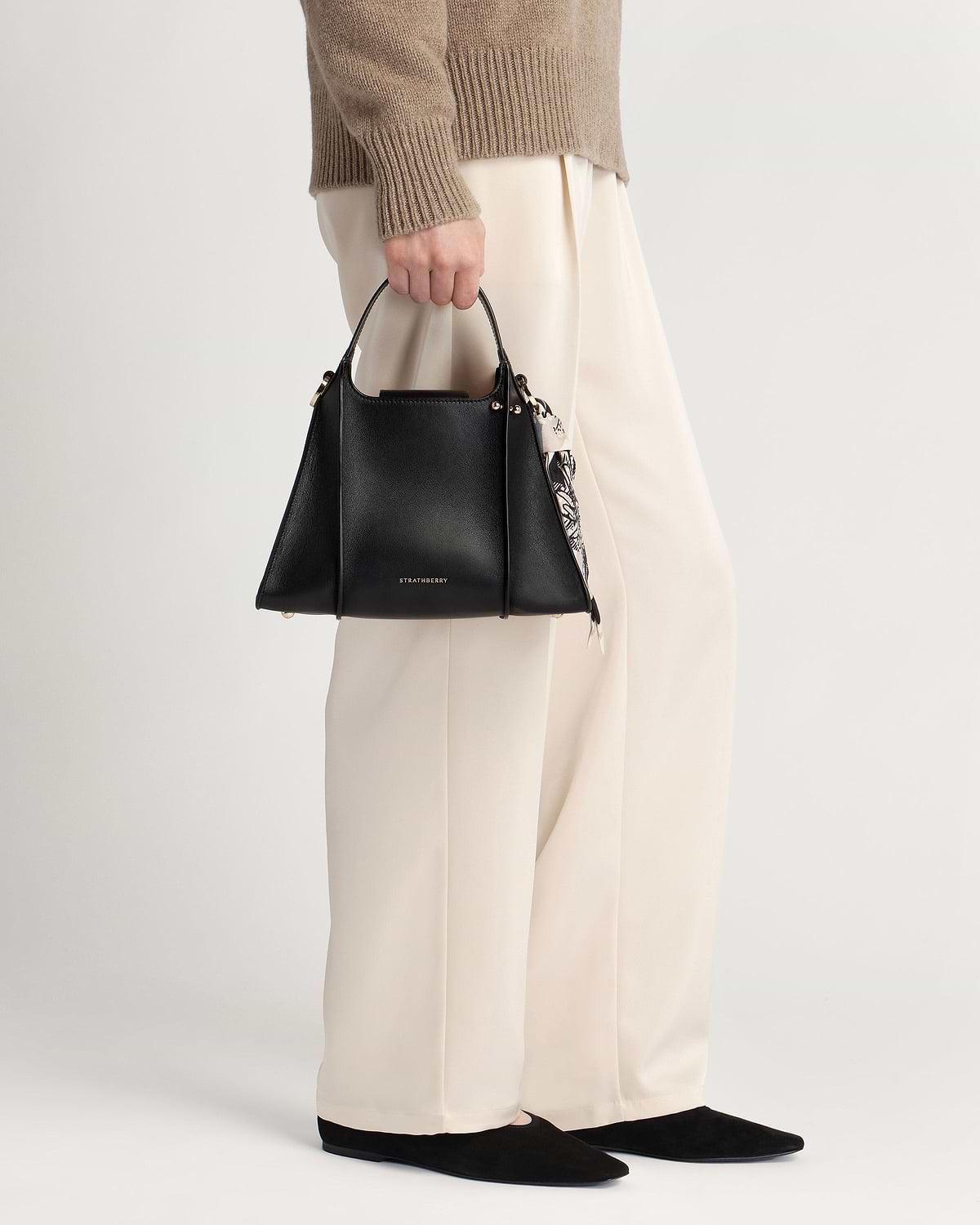 Model holding black Strathberry Arkshell handbag with patterned scarf, cream trousers.