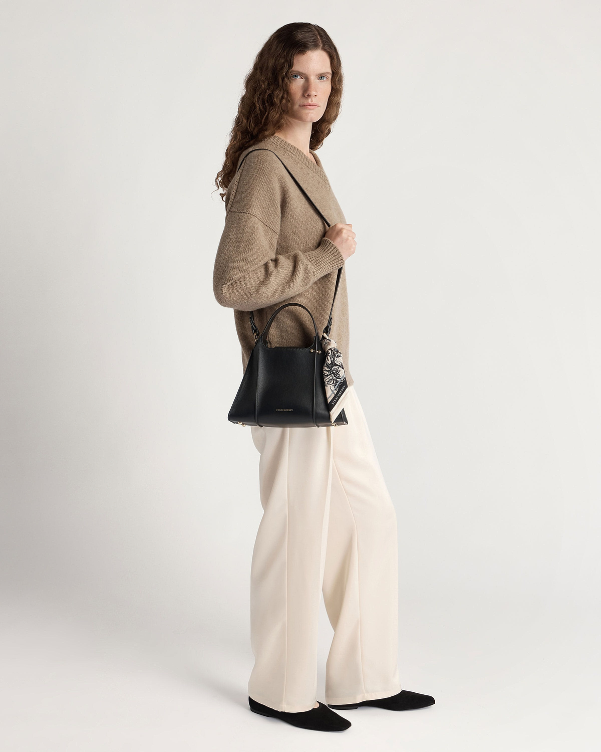 Woman wearing beige sweater and cream trousers carrying black Arkshell bag with patterned scarf.