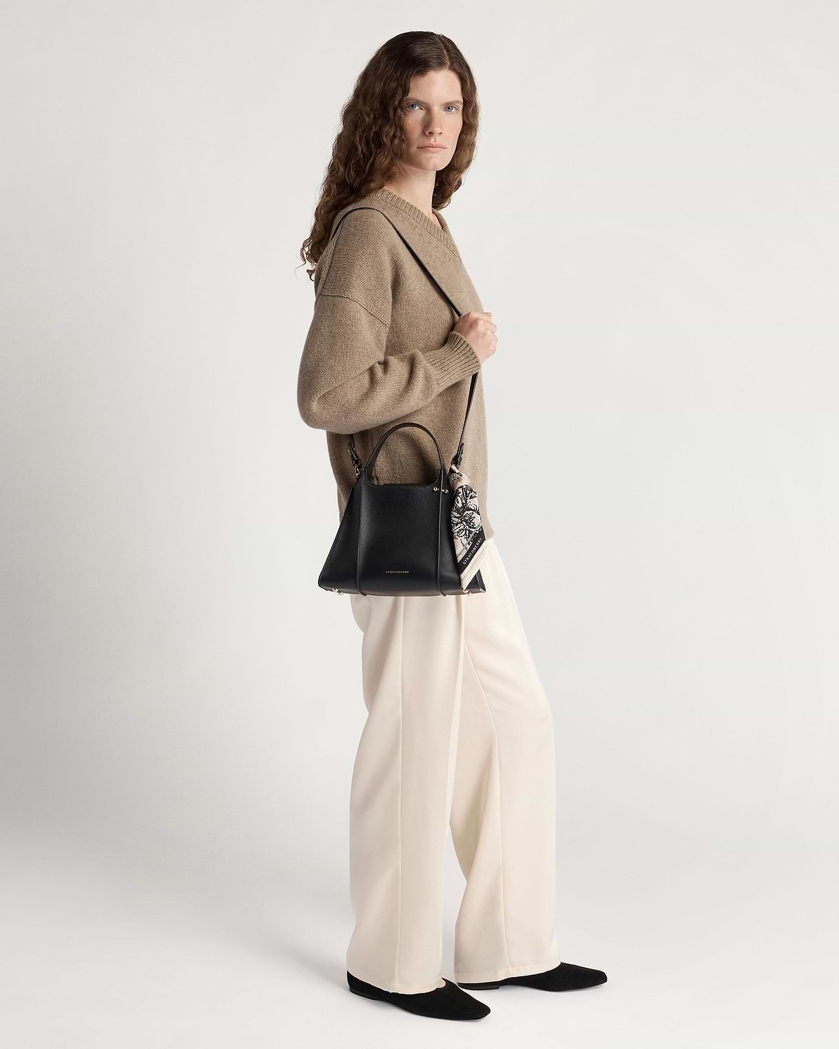 Woman wearing beige sweater and cream trousers carrying black Arkshell bag with patterned scarf.