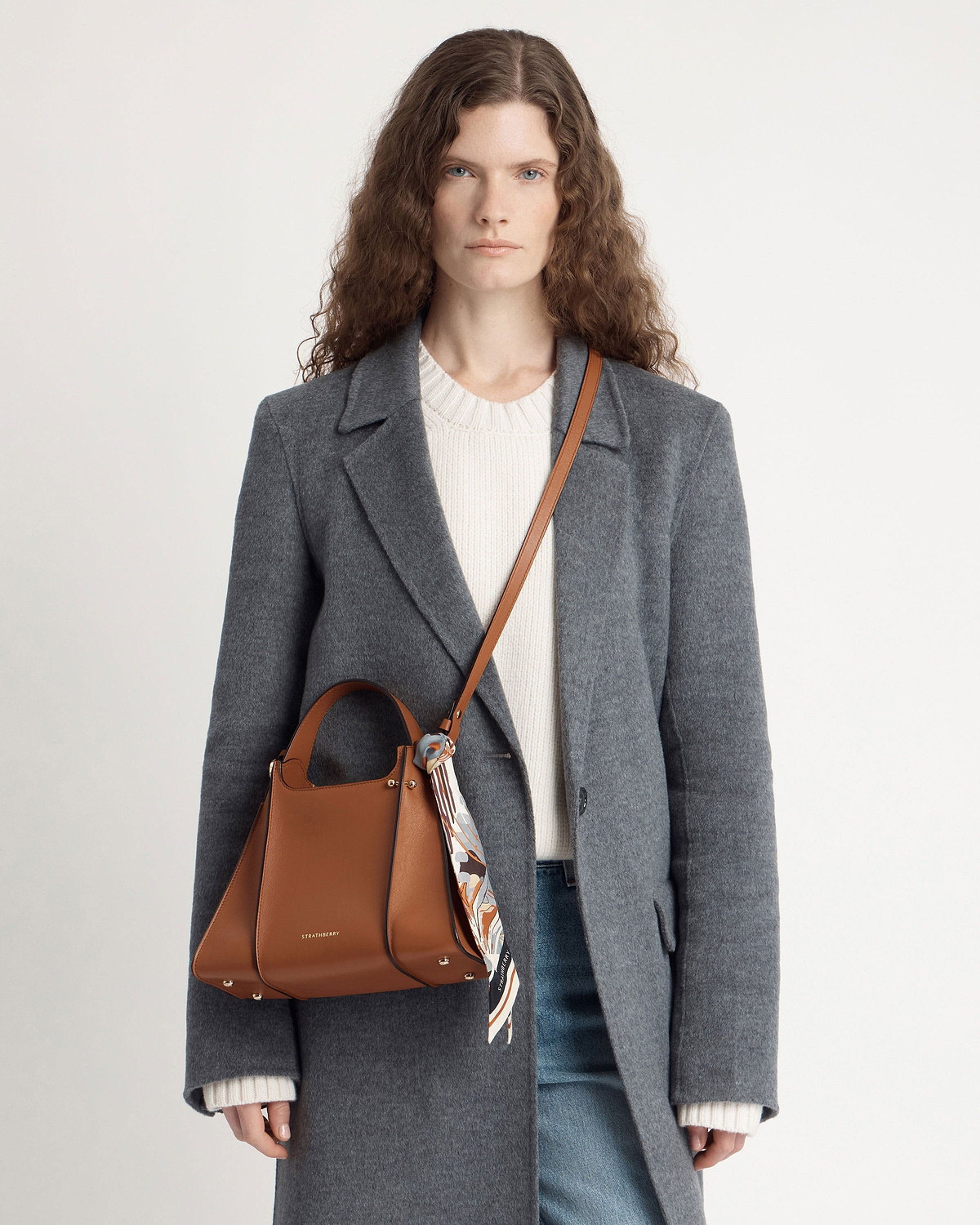 Woman in grey coat wearing tan Strathberry Arkshell bag with patterned scarf.
