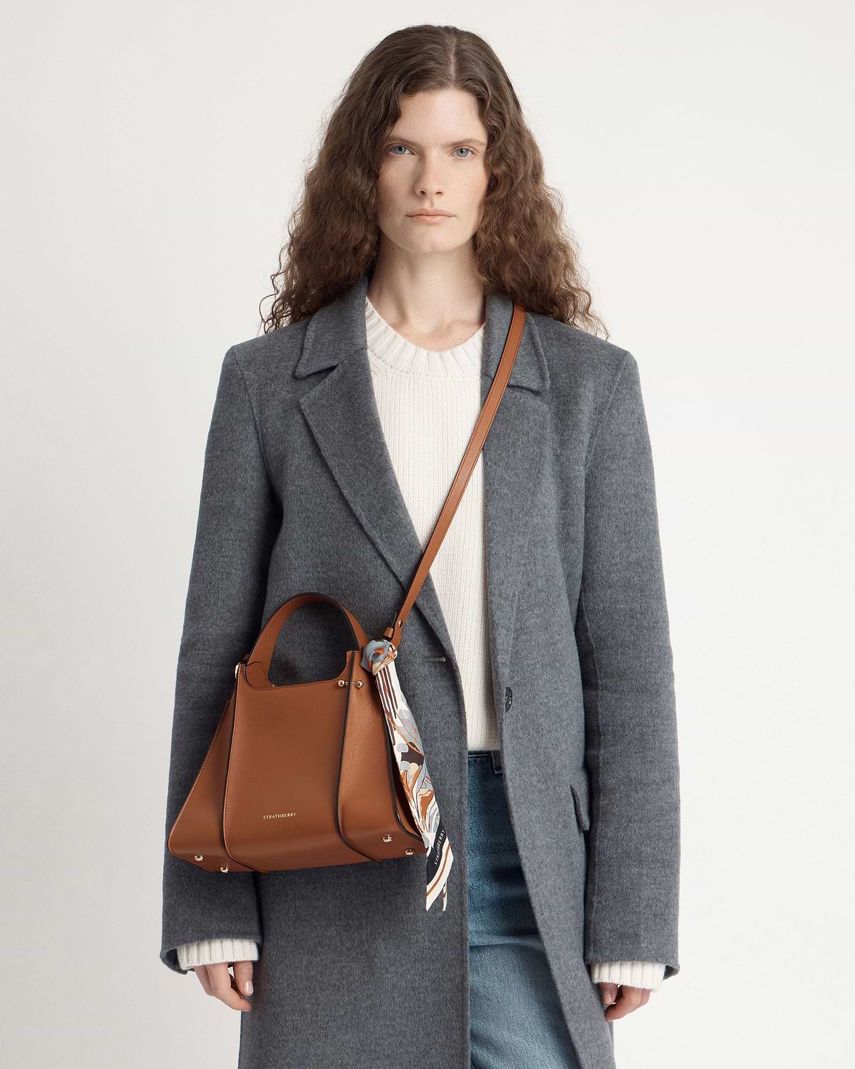 Woman in grey coat wearing tan Strathberry Arkshell bag with patterned scarf.