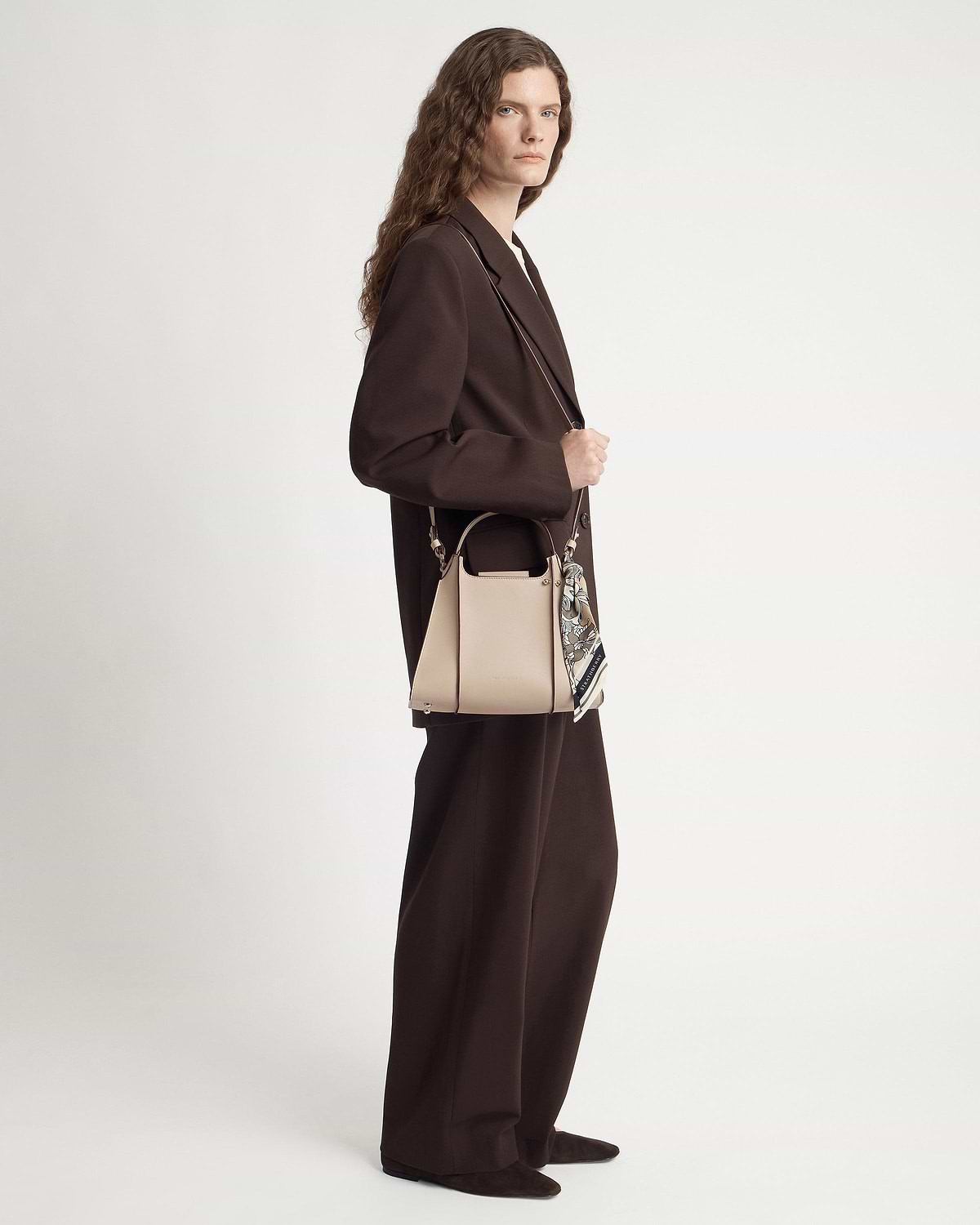 Model in brown suit holding an oat-coloured structured leather handbag with silk scarf.