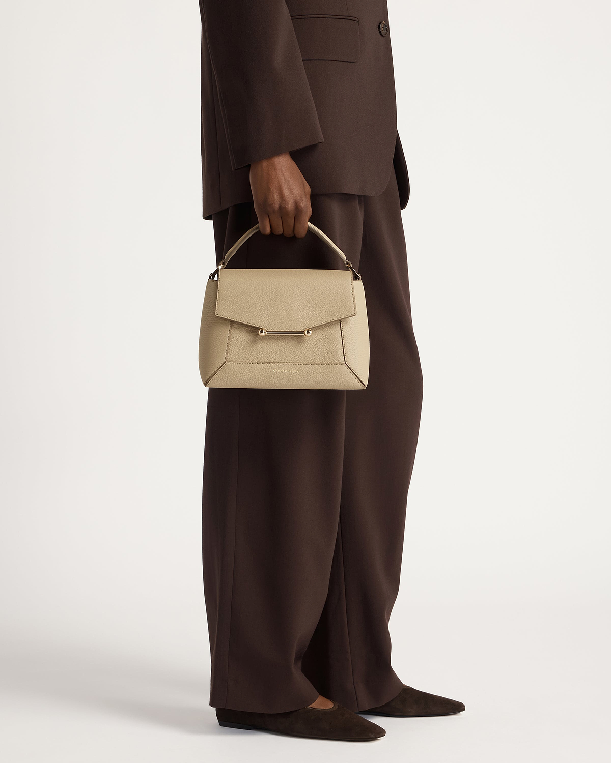 Model holding a sand mosaic top-handle bag against a brown tailored suit.