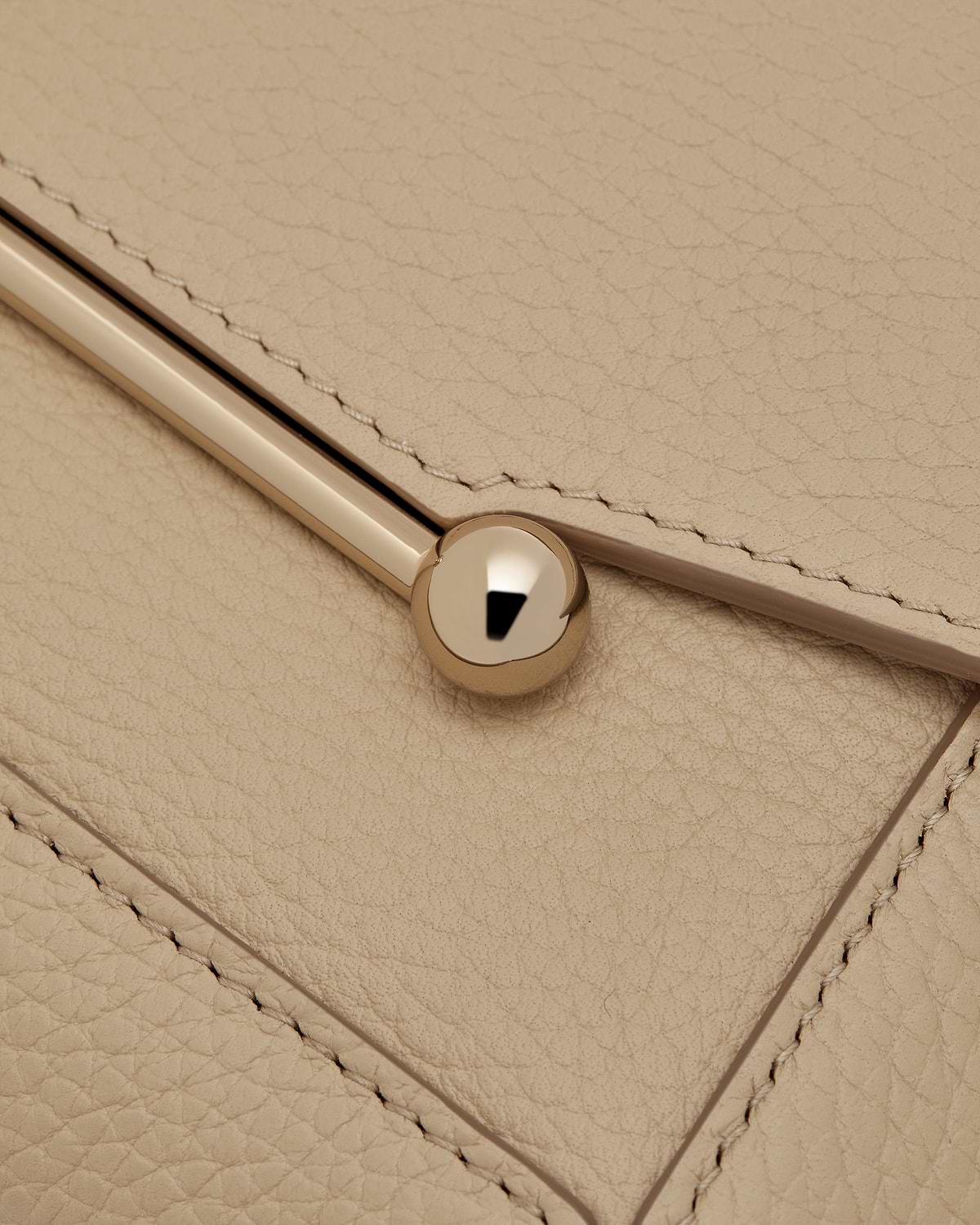 Close up of sand leather flap showing stitching, a gold bar and round stud.