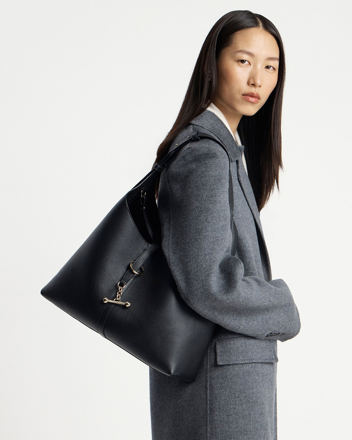 Model wearing a grey coat carrying a black hobo shoulder bag with gold hardware.