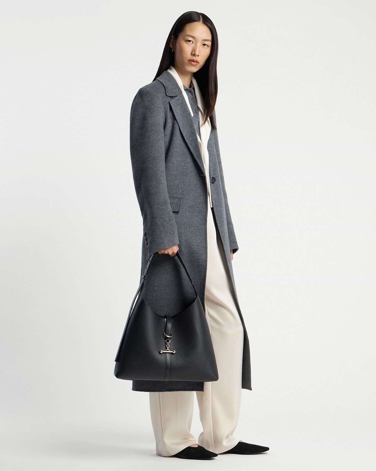 Model wearing grey coat holding black leather hobo bag with gold hardware.