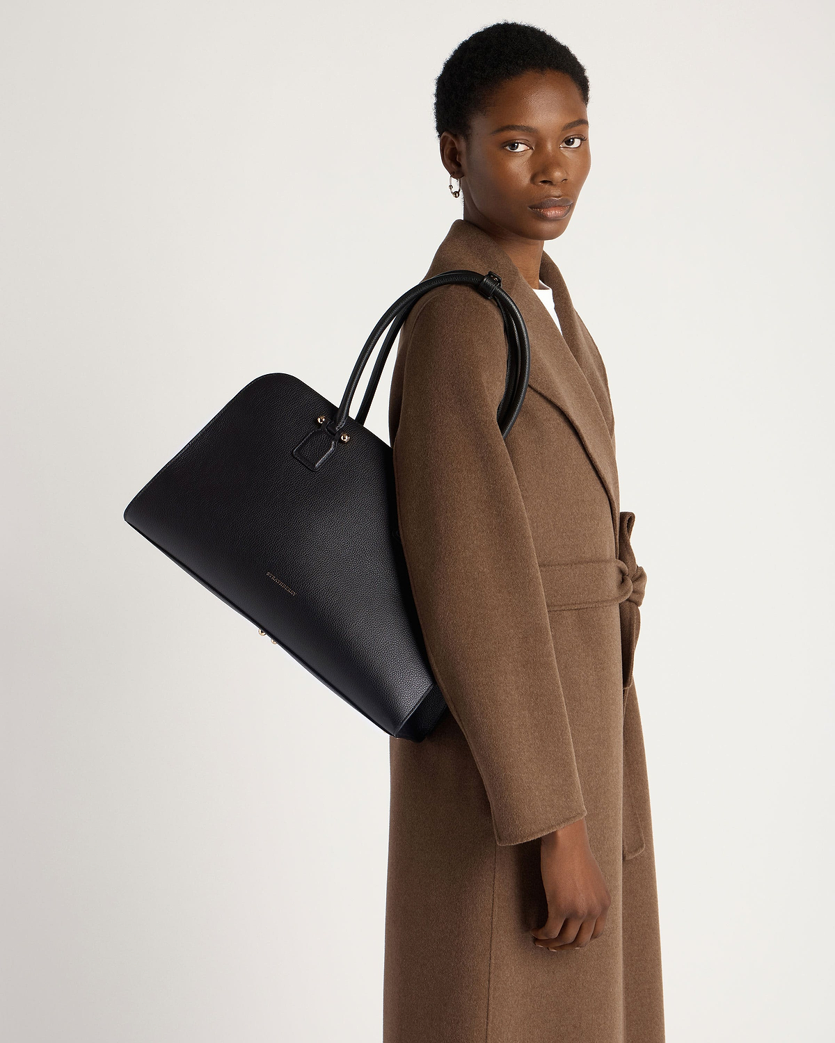 Model in brown coat carrying black Strathberry Georgia Maxi leather tote on shoulder